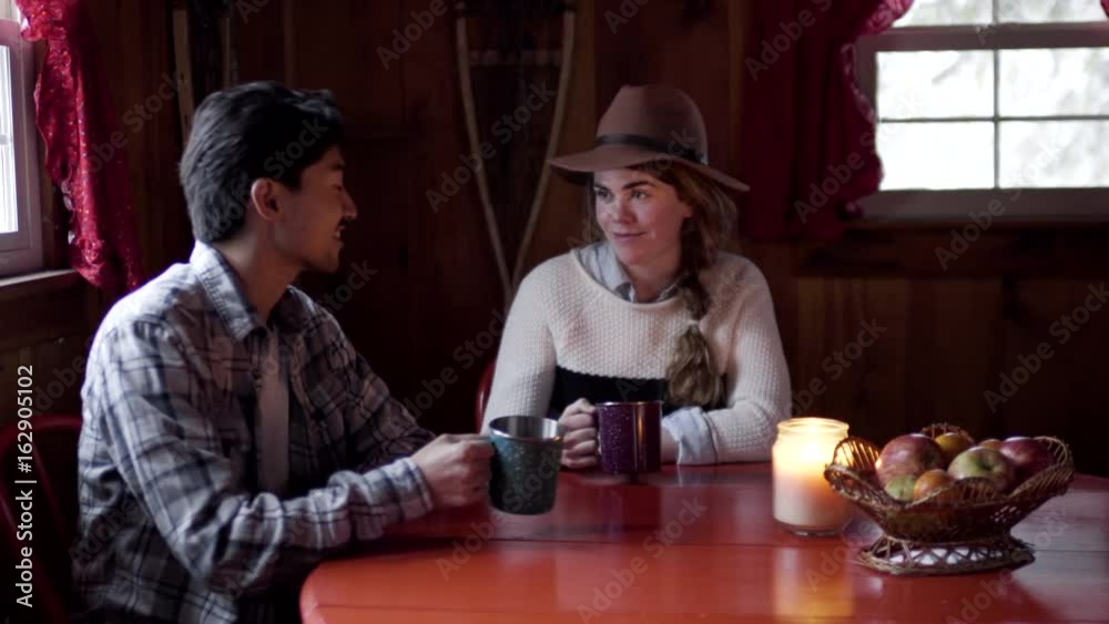 Cute Couple Sit At Kitchen Table In Cozy Cabin, They Cheers With Enamel Mugs Cute Couple Sit At Kitchen Table In Cozy Cabin, They Cheers With Enamel Mugs