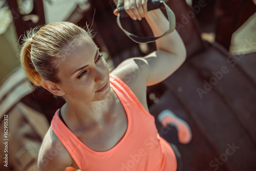 Close-up portrait of an athlete in uniform during exercise with TRX exercise outdoors. Beautiful young woman in an orange T-shirt.