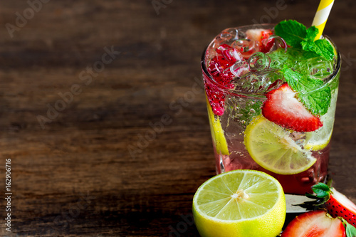 Strawberry lemon lime mojito in clear glass on rustic wood table. Homemade beverage strawberry soda ingredients with strawberry, lemon or lime and mint leaf. Homemade infused water with copy space.