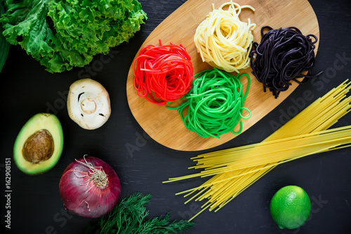 Multicolored pasta on wooden board and raw vegetables on dark table. Flat lay. Top view.