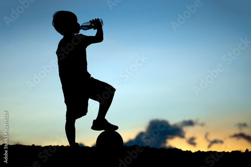 Silhouette of little boy drinking water from a bottle at sunset with the sun in the background