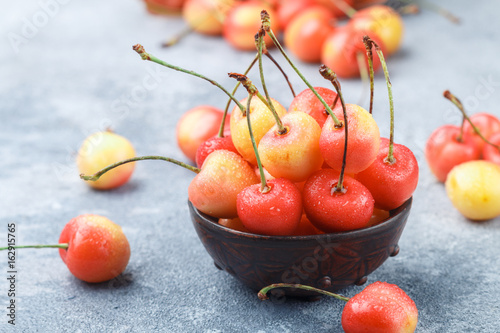 Red and yellow Rainier cherries with drops of water in a clay bowl on the gray concrete surface. Selective focus photo