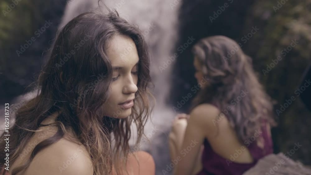 Bohemian Models Sit On Cliff Across From Waterfall, One Looks At Camera, Poses Bohemian Models Sit On Cliff Across From Waterfall, One Looks At Camera, Poses