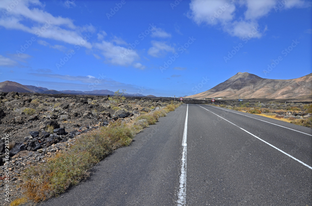An empty road through volcanic landscape of Lanzarote, the Canary Islands. Volcanoes in the background and blue sky with picturesque clouds.