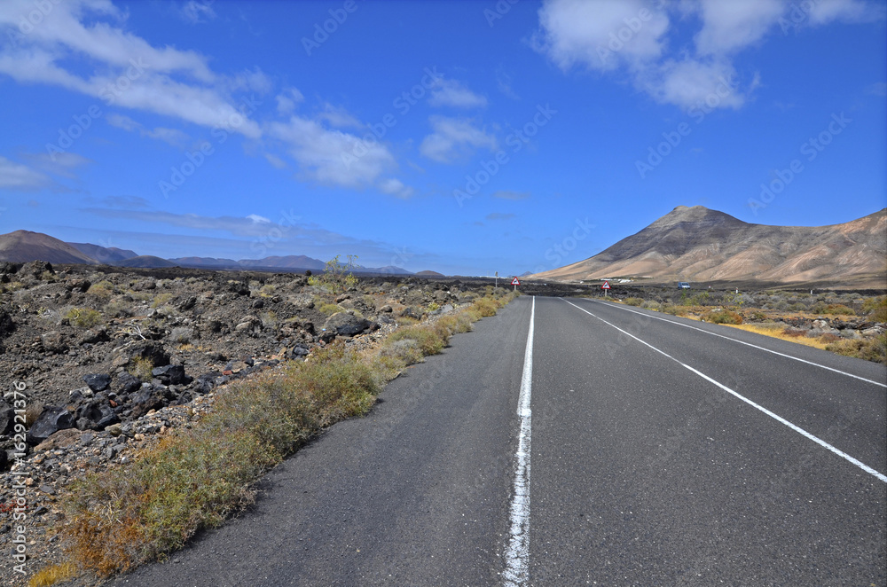 An empty road through volcanic landscape of Lanzarote, the Canary Islands. Volcanoes in the background and blue sky with picturesque clouds. An empty road through volcanic landscape of Lanzarote, the Canary Islands. Volcanoes in the background and blue sky with picturesque clouds.