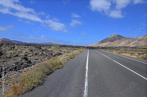 An empty road through volcanic landscape of Lanzarote, the Canary Islands. Volcanoes in the background and blue sky with picturesque clouds.