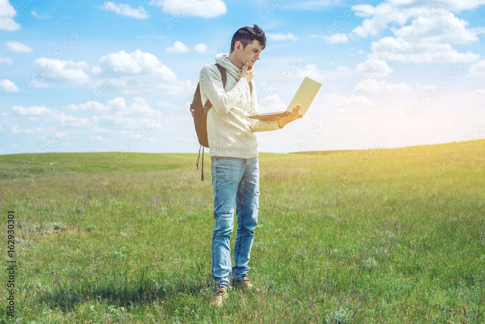 Young man sitting on a green meadow with laptop wireless on the background of blue cloudy sky