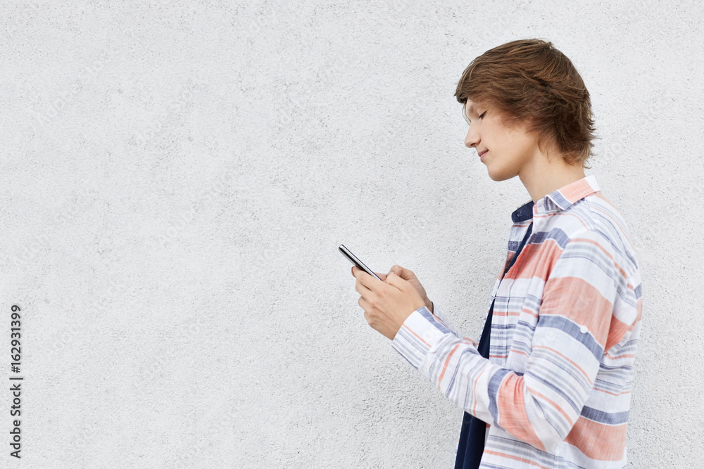 Stylish teenage boy standing sideways holding cell phone isolated over white concrete wall with copy space for your advertisment or promotional text. People, modern technologies and lifestyle Stylish teenage boy standing sideways holding cell phone isolated over white concrete wall with copy space for your advertisment or promotional text. People, modern technologies and lifestyle