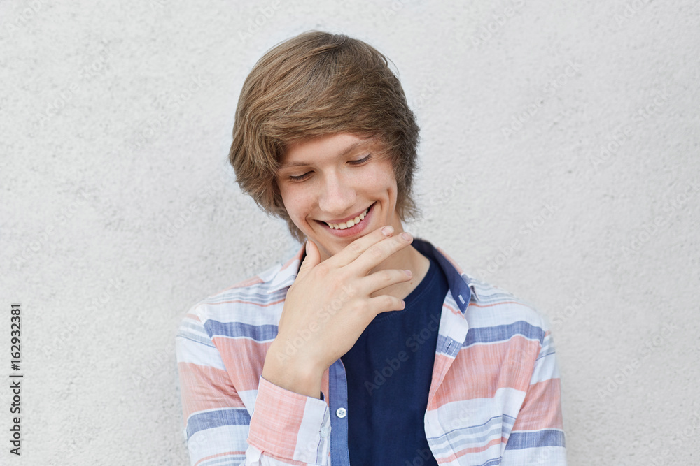 Portrait of smiling shy boy with trendy hairdo wearing casual shirt looking  down holding hand on