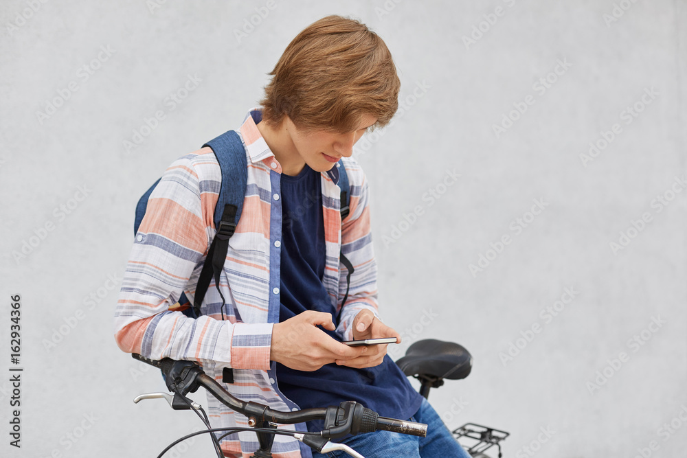 Portrait of hipster guy with trendy hairstyle wearing shirt and jeans holding backpack standing near his bicycle using smart phone looking attentively playing games. People and technology concept Portrait of hipster guy with trendy hairstyle wearing shirt and jeans holding backpack standing near his bicycle using smart phone looking attentively playing games. People and technology concept