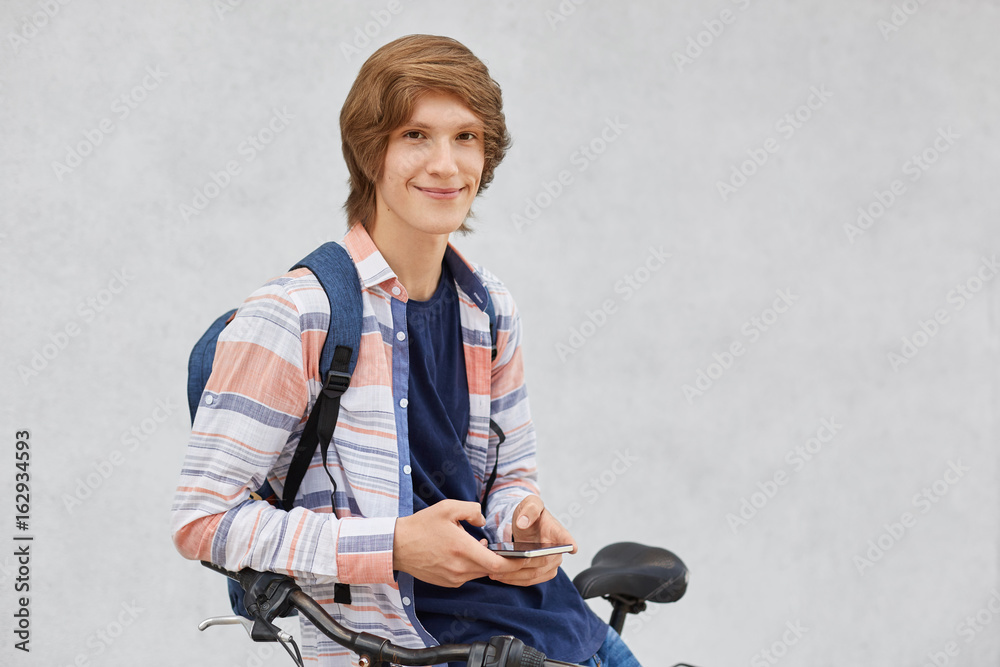 Handsome hipster guy with trendy haircut wearing shirt and jeans holding backpack sitting on bicycle holding cell phone looking with smile into camera. People, teenagers, modern technologies concept Handsome hipster guy with trendy haircut wearing shirt and jeans holding backpack sitting on bicycle holding cell phone looking with smile into camera. People, teenagers, modern technologies concept