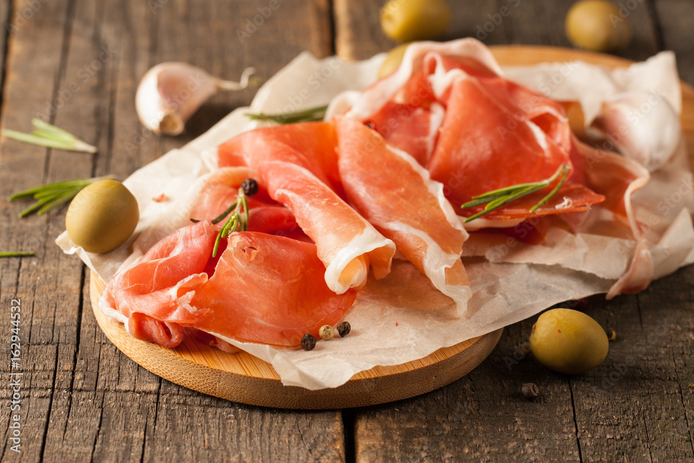 closeup of thin slices of prosciutto with mixed olives and paprika on wooden cutting board