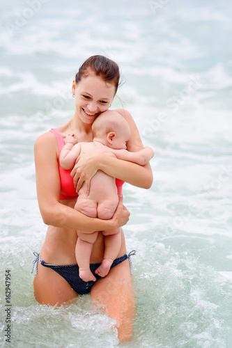 A young beautiful mother is holding a baby in her arms. This is his first bathing in the sea.