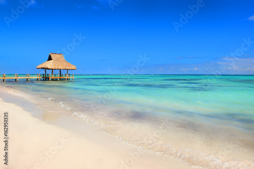 Tropical white sandy beach. Palm leaf roofed wooden pier with gazebo on the beach. Punta Cana, Dominican Republic © alekosa