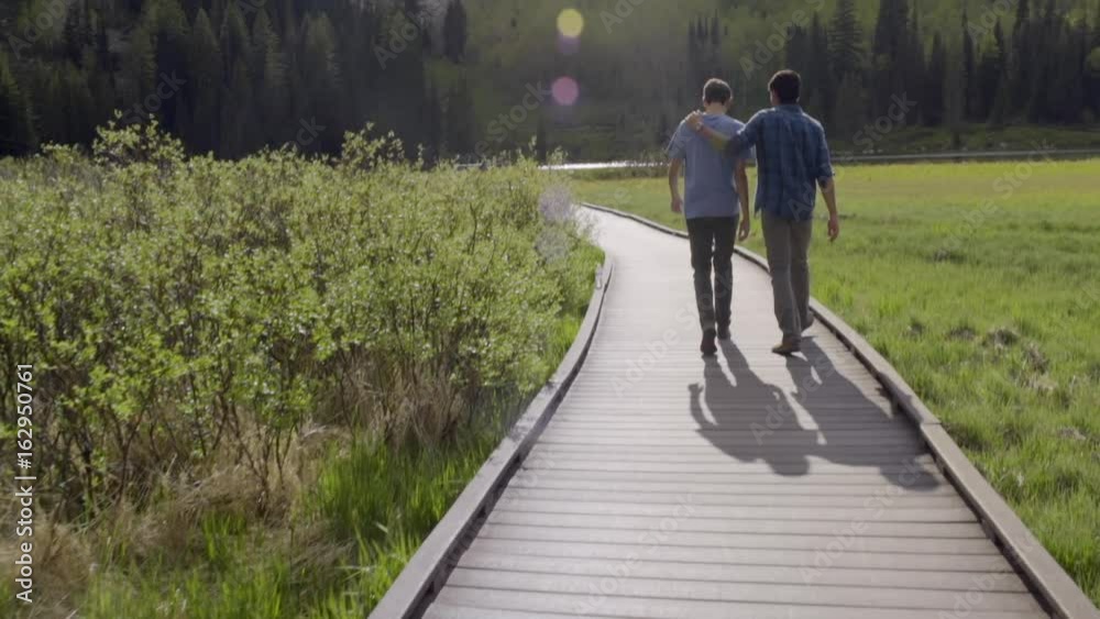 Best Friends Walk Down Boardwalk Toward Mountain Lake, Boy Puts His Hand On His Friend's Back Best Friends Walk Down Boardwalk Toward Mountain Lake, Boy Puts His Hand On His Friend's Back