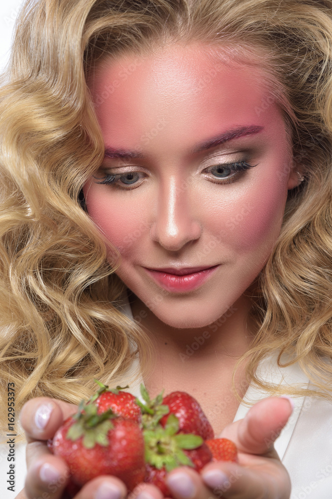 Beautiful blonde woman face with pink makeup. Glamour fashion model portrait with strawberry. Colorful pigment on the face, glossy skin, bright make-up. White backdrop Beautiful blonde woman face with pink makeup. Glamour fashion model portrait with strawberry. Colorful pigment on the face, glossy skin, bright make-up. White backdrop