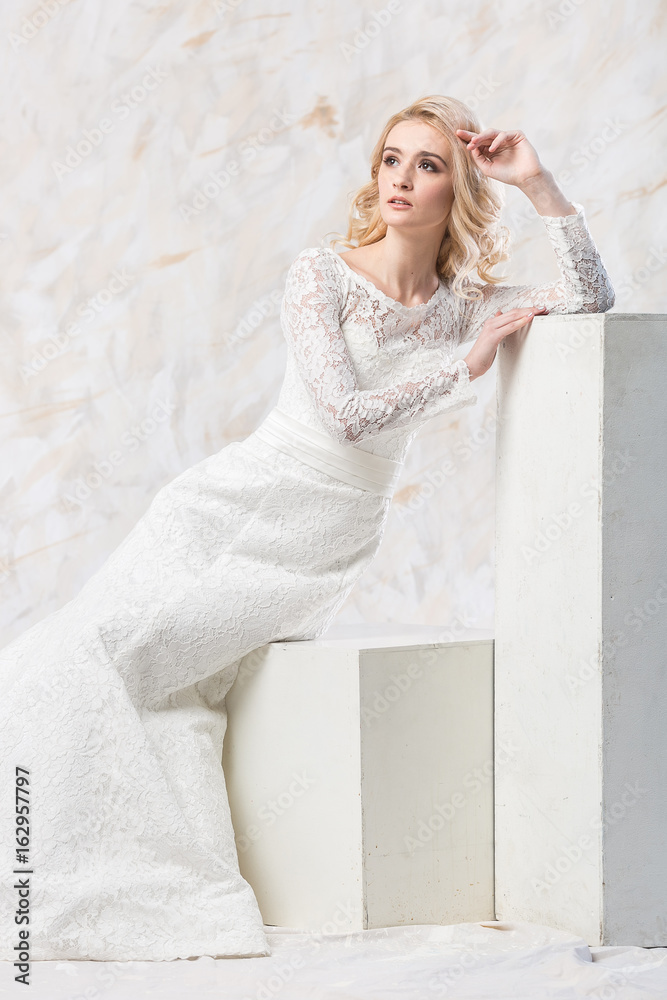 Fototapeta premium fashionable dress, beautiful blonde model, bride hairstyle and makeup concept - luxury young woman in white wedding gown sitting indoors on light background, female posing in the studio