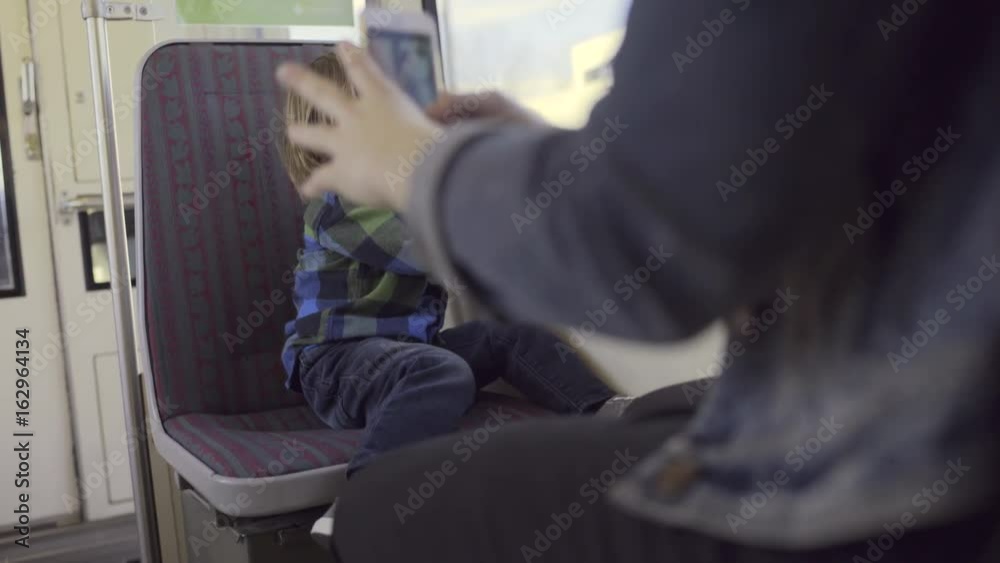 Little Boy Plays With His Dinosaur Toy On The Train, His Mother Takes A Photo Of Him With Her Smart Phone Little Boy Plays With His Dinosaur Toy On The Train, His Mother Takes A Photo Of Him With Her Smart Phone