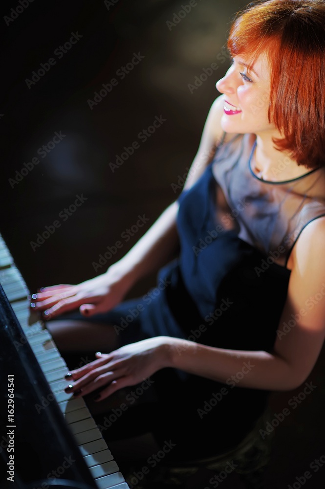 Fototapeta premium Beautiful cheerful girl, illuminated by a beam of bright light, sits at the piano and smiles genuinely on a dark background closeup