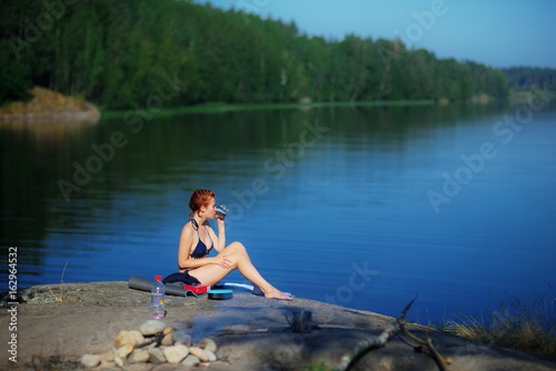 Beautiful young red-haired girl in a swimsuit drinking tea, sitting on the stone above the river, enjoying the peace, tranquility and beauty of nature and sunlight