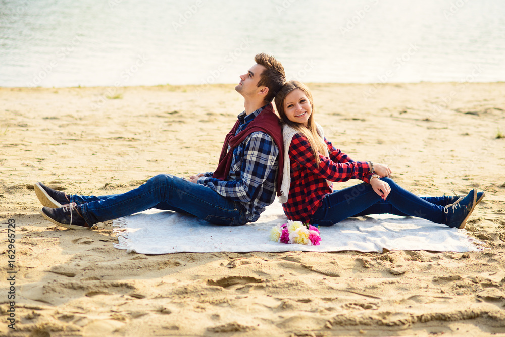 Stylish young couple in shirts and jeans while walking on the shore of a lake. Beautiful attractive girl and her handsome boyfriend leans to each other sitting on a plaid.