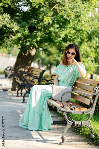 Young beautiful woman in a light green pastel long dress is sitting on a park bench.