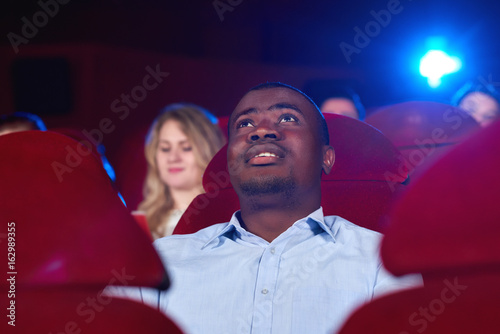 Slika na platnu Low angle shot of a young African man watching a movie sitting at the cinema auditorium fascinated interested viewer spectator entertaining activity person concept