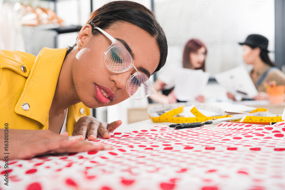 Fototapeta premium young african american dressmaker fitting polka dot fabric while her colleagues sitting on background