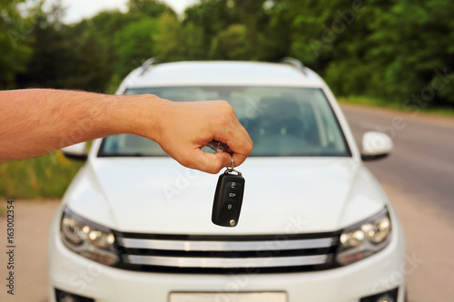 Male hand holding car keys on blurred background with new white car, green tree and road in summer outdoors.