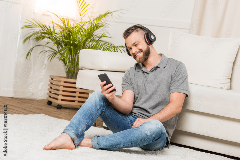 Cheerful bearded young man in headphones using smartphone and listening music at home