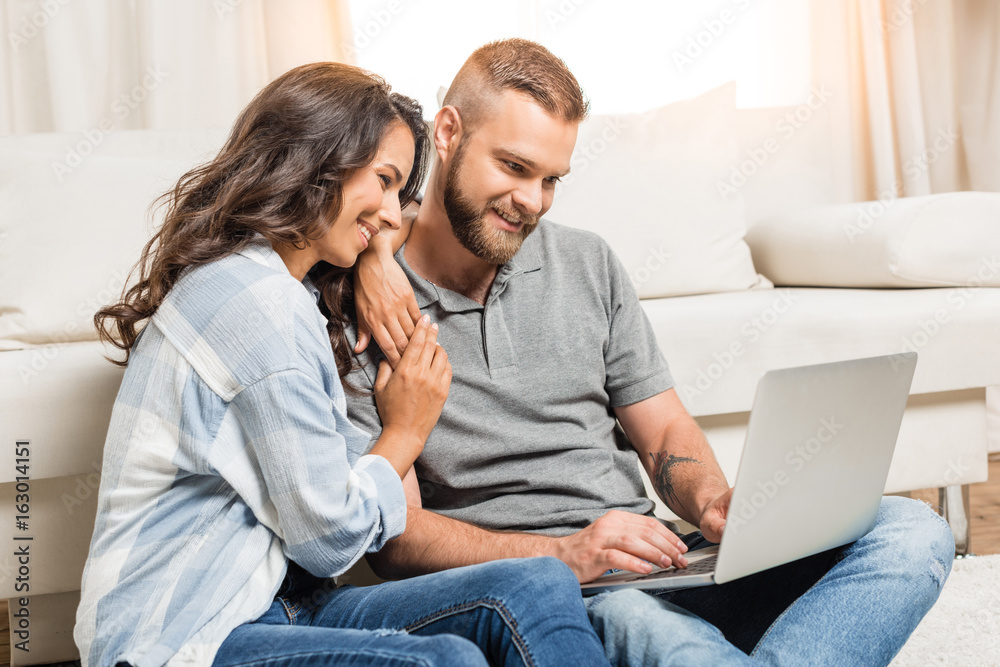 young happy couple using laptop while sitting on carpet at home