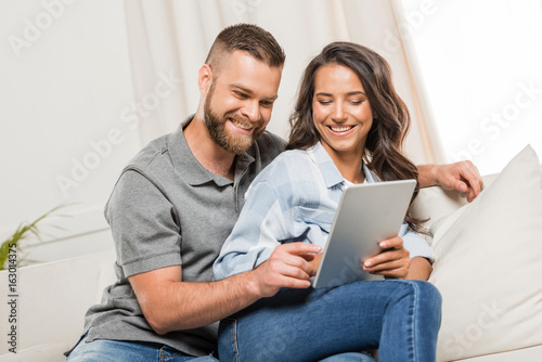 young smiling couple using digital tablet while sitting on sofa at home