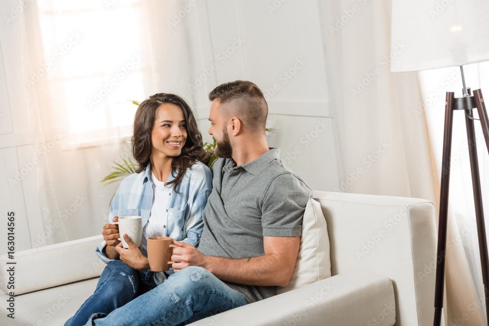 happy couple drinking coffee and looking at each other while sitting on sofa at home happy couple drinking coffee and looking at each other while sitting on sofa at home
