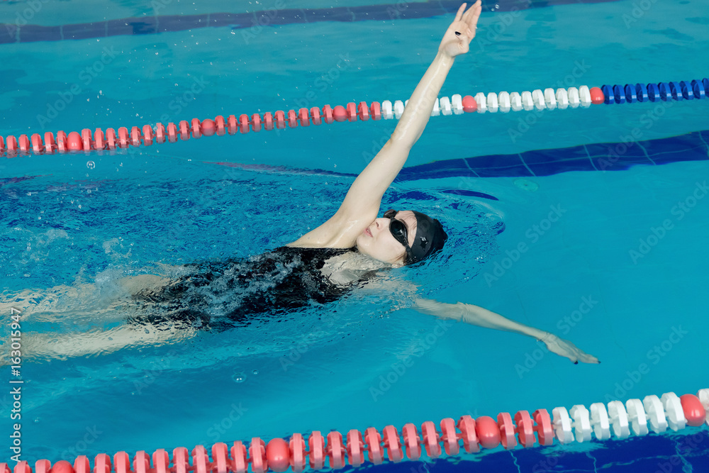 Young girl in goggles and cap swimming backstroke