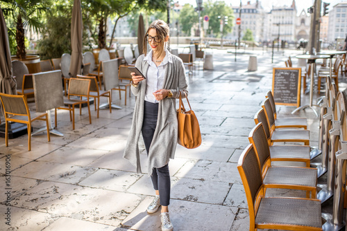 Photography Young stylish businesswoman walking with phone and bag on the street with cafes