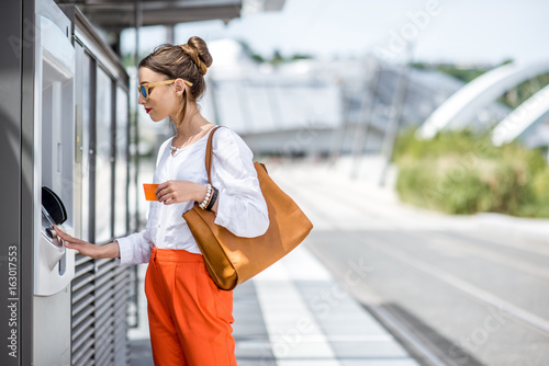 Young woman buying a ticket for public transport at the automatic machine or withdrawing money with a card standing outdoors at the modern city photo