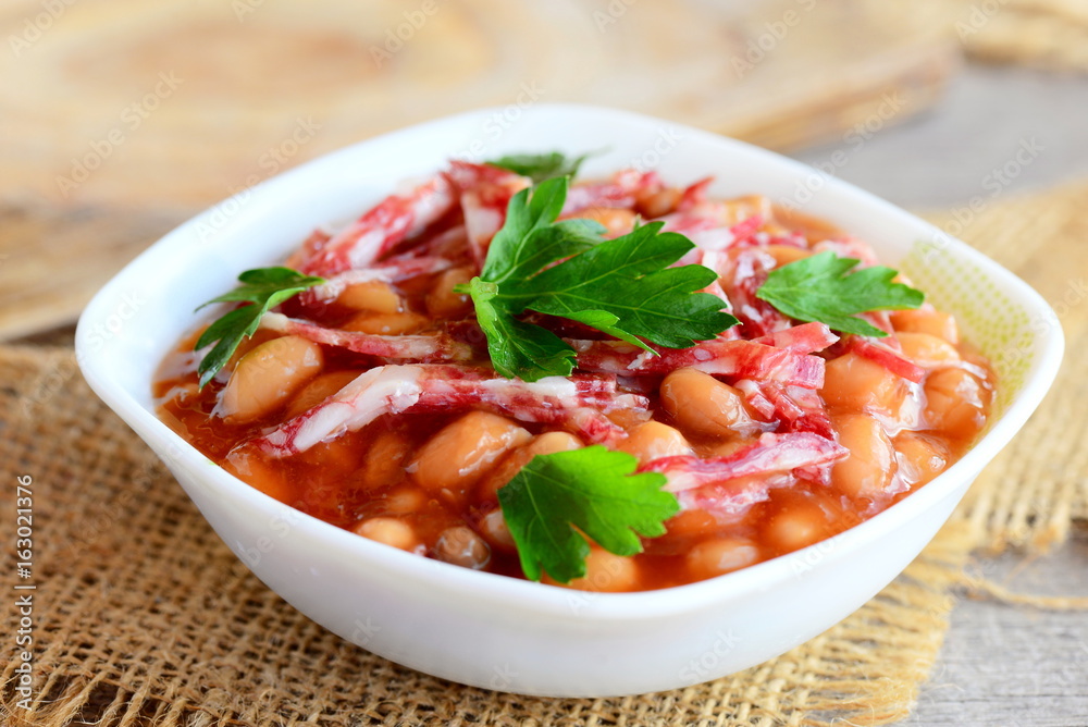 Smoked sausages with white beans in tomato sauce. Simple white bean and smoked sausage stew in a white bowl and on a wooden old table. Home nutritious stew. Closeup Smoked sausages with white beans in tomato sauce. Simple white bean and smoked sausage stew in a white bowl and on a wooden old table. Home nutritious stew. Closeup