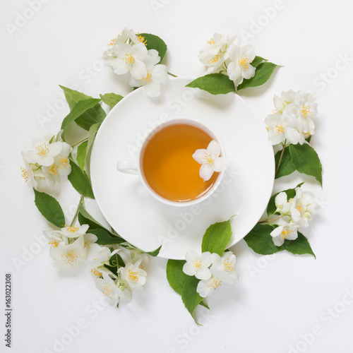 Jasmine leaves and flowers around cup of green tea on white background. Top view and concept.