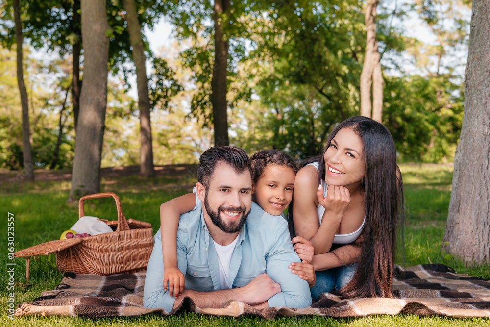 happy family looking at camera while lying on blanket on picnic in park