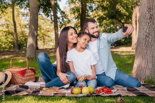 Fototapeta Naklejka Na Ścianę i Meble -  happy family taking selfie on smartphone while having picnic in park