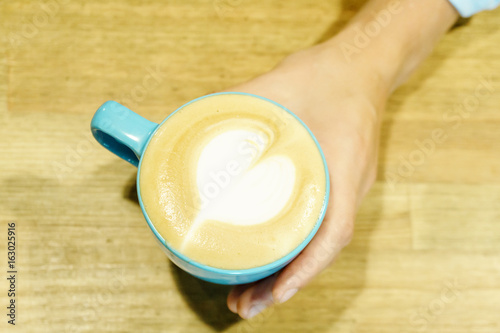 Woman hand holding a blue cup of cappuccino on wooden table background. Heart milk froth.
