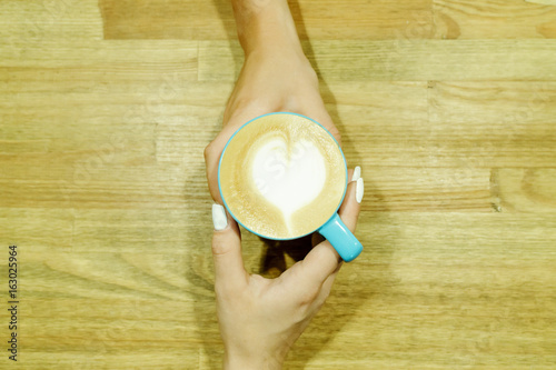 Cappuccino with heart froth between two hands on wooden table. Human pastime