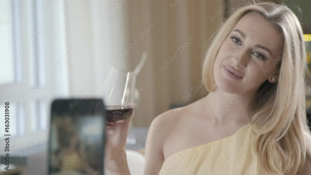 Photo by phone: the girl is drinking wine. Gentle and pretty woman in a dress drinks wine, at which time a man takes a photo on a smartphone. Beautiful woman drinks wine, she is photographed on phone