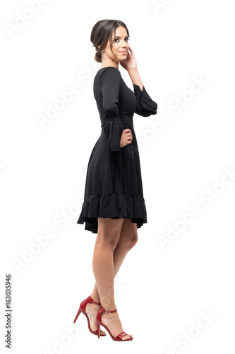 Side view of tanned Hispanic woman in black dress posing at camera touching hair. Full body length portrait isolated on white studio background. 