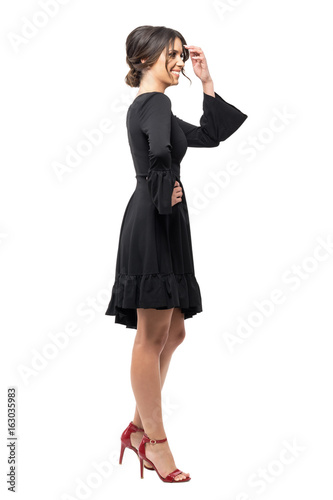 Laughing young Latin woman in black dress looking away. Side view. Full body length portrait isolated on white studio background. 