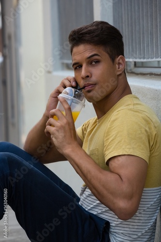 Young man drinking juice while talking on smart phone by wall