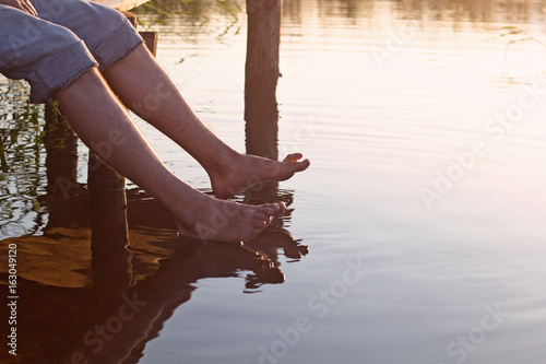 a man sits  on a wooden bridge and dip feet in warm water for a lake in the summer