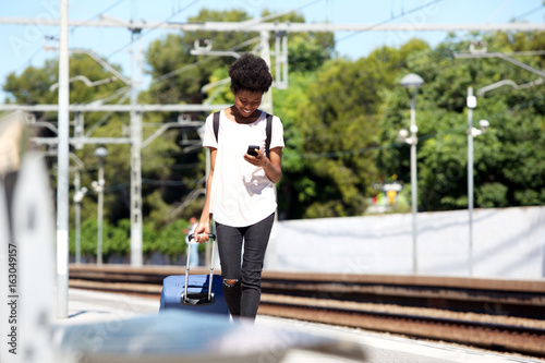 Young african woman walking on railway station with suitcase and mobile phone