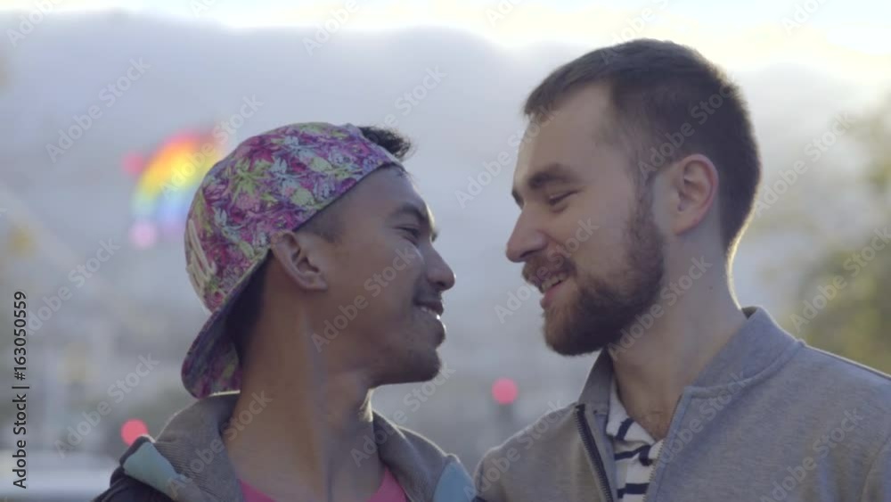 Vidéo Stock Interracial Gay Couple Kiss And Laugh, Gay Pride Flag Blows In Breeze Behind Them | Adobe Stock