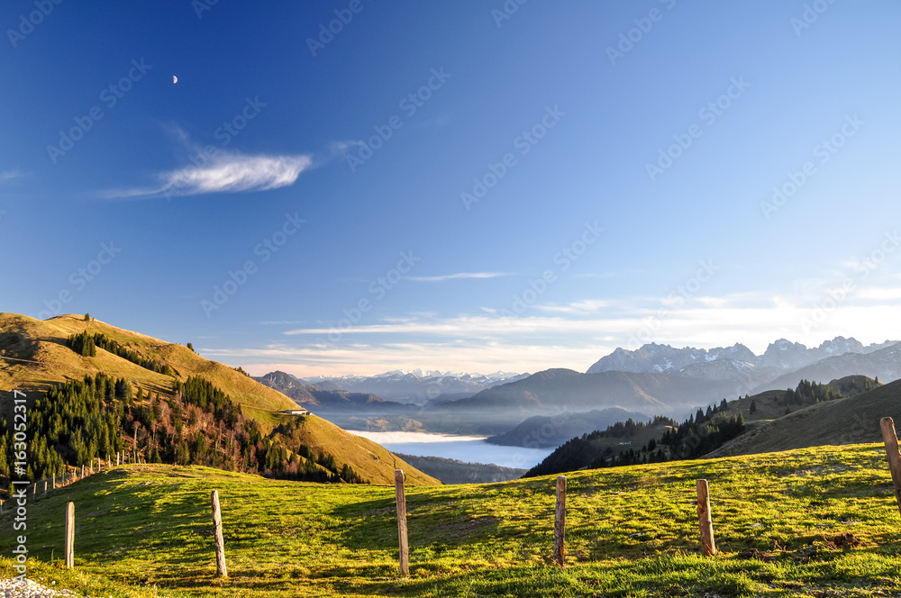 Beautiful evening view near Wandberg mountain in the Chiemgau Alps mountain range in Austria. Tranquil scene with a fence, a fresh lawn and mountains in the background and fog in the lowland. 